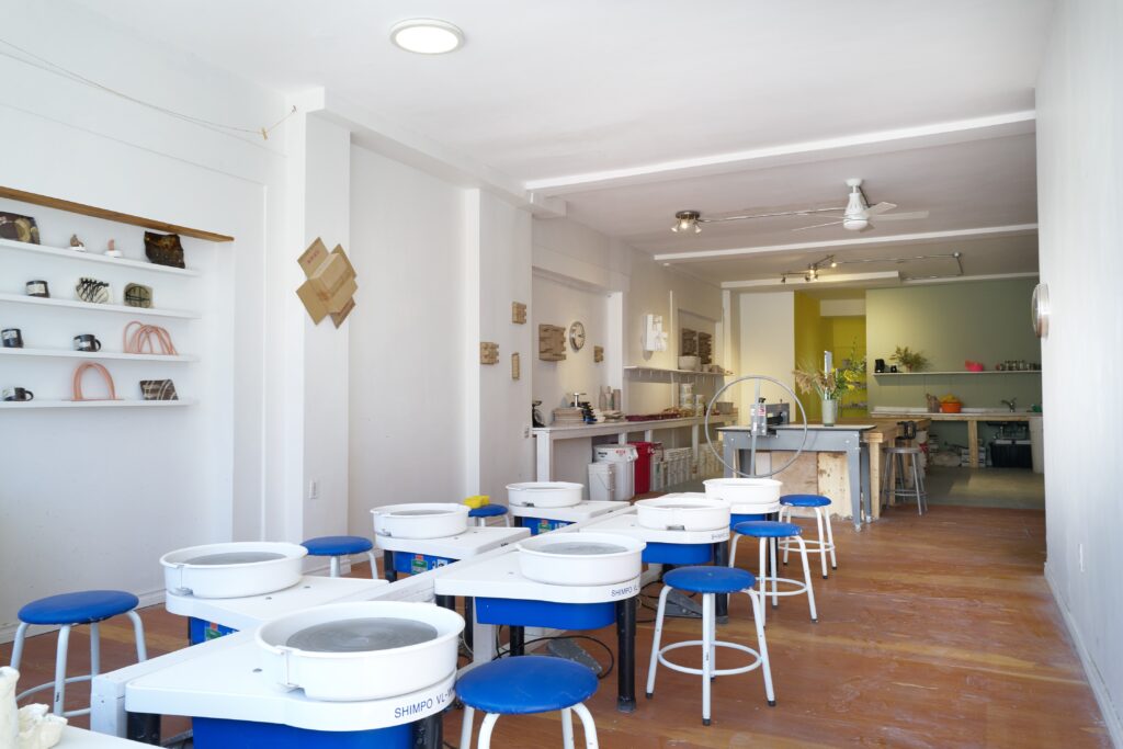 Spacious pottery studio with blue stools and white pottery wheels. Shelves display ceramics, and a cozy workspace with a warm atmosphere is visible in the back.
